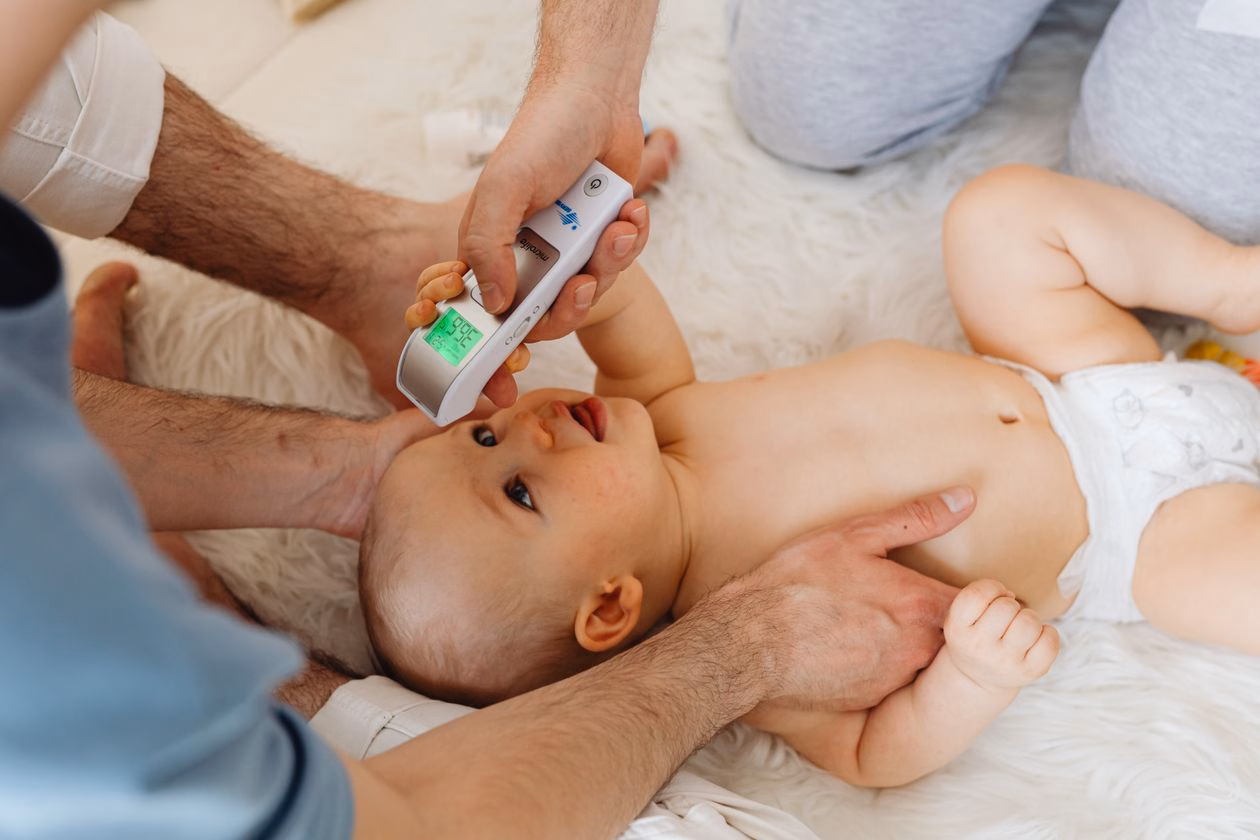 Baby thermometer in India: father holding a digital thermometer against a baby lying on white bedding for a quick home temperature check