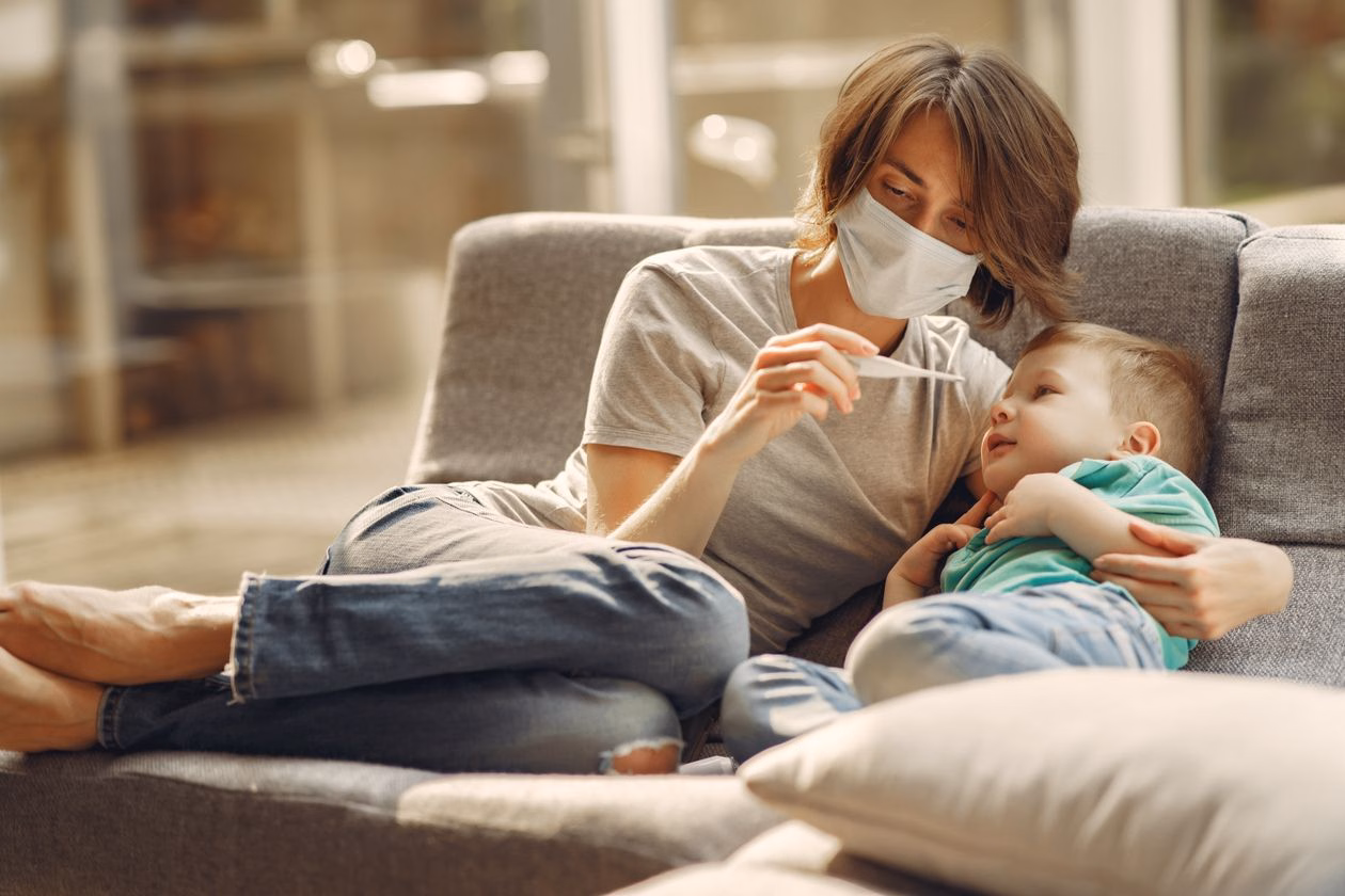 Mother checking her child's temperature with a thermometer while they rest together on a sofa, a calm everyday home routine